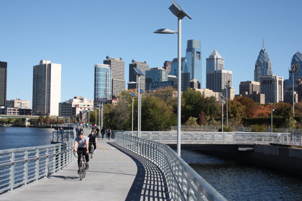 Bicyclists riding their bikes on a trail above the Schuylkill River with a view of the Center City skyline