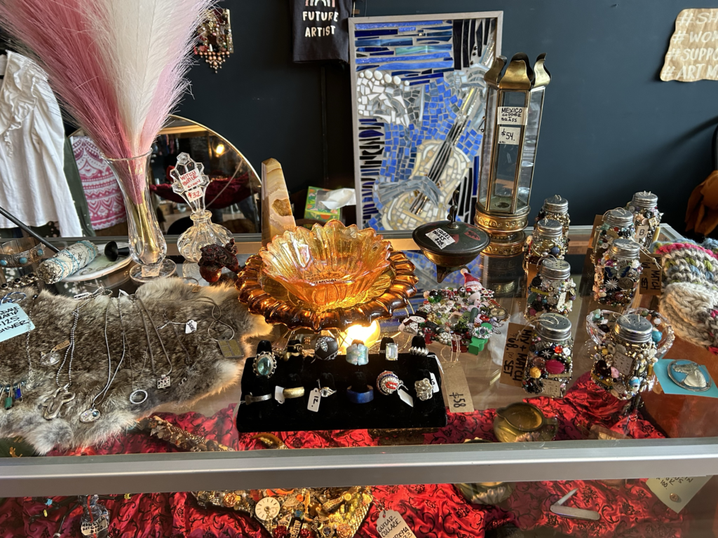 A counter at a small shop with items on it such as necklaces, rings, dishes, crystal, and jars of beads.