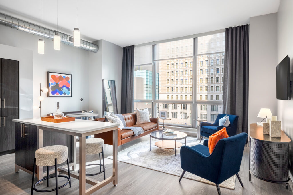 Living area in The Collins at Midtown. A bright living room, an island table with two stools, a couch, and two chairs in front of a large window