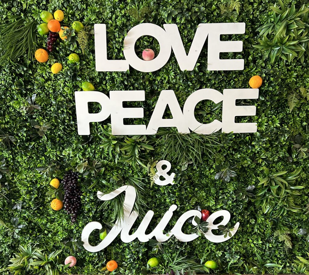 A green wall covered in artificial plants and fruit with white words on top that read "Love, Peace, & Juice"