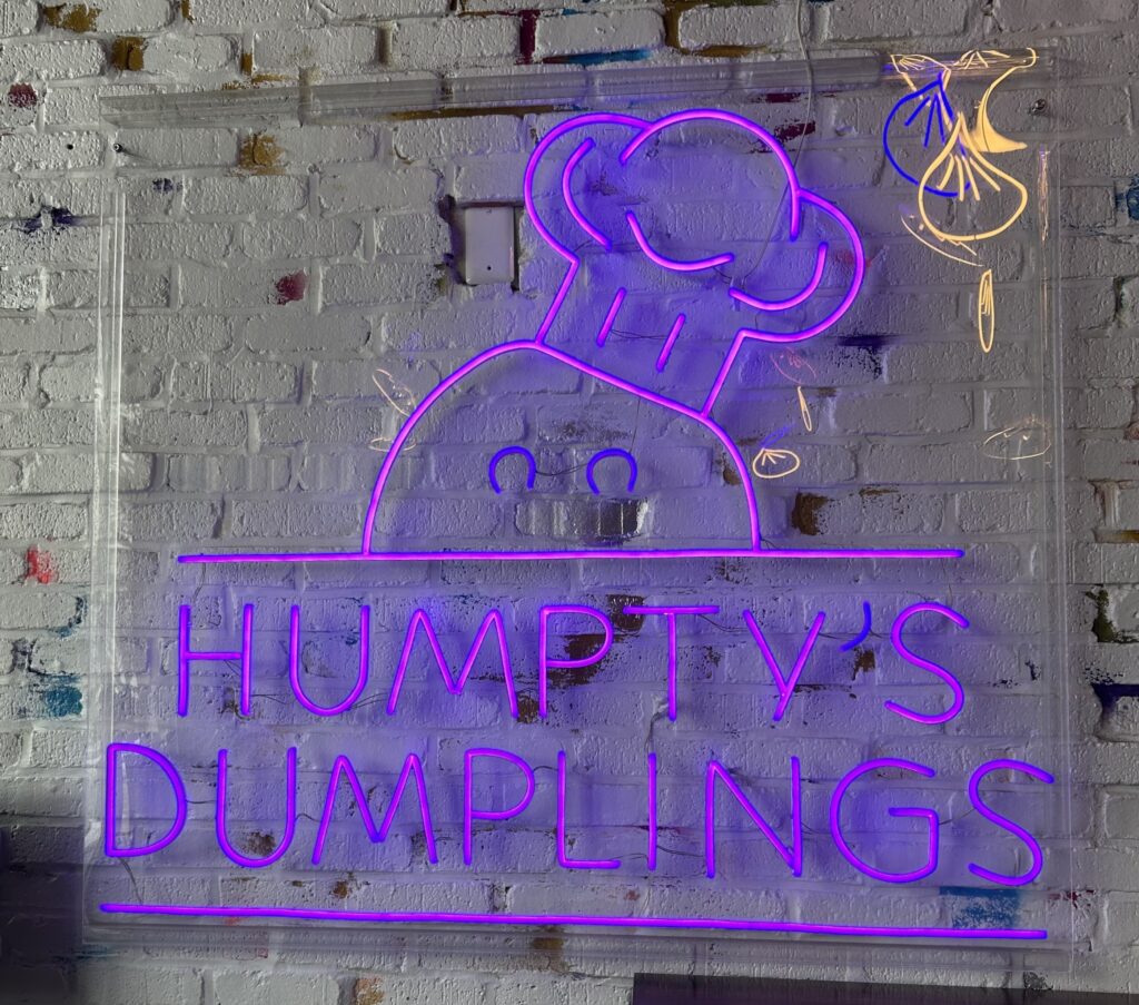 A white brick wall with a purple neon sign that reads "Humpty's Dumplings" with the logo of a dumpling in a chef's hat