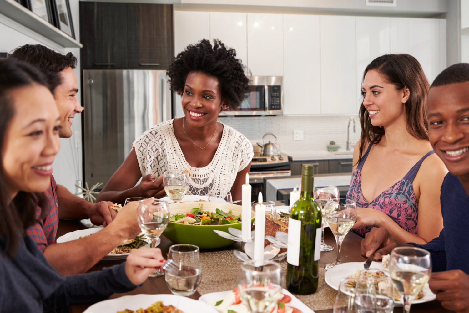 Six friends sitting around a dinner table eating salad, drinking wine, and laughing in an apartment kitchen