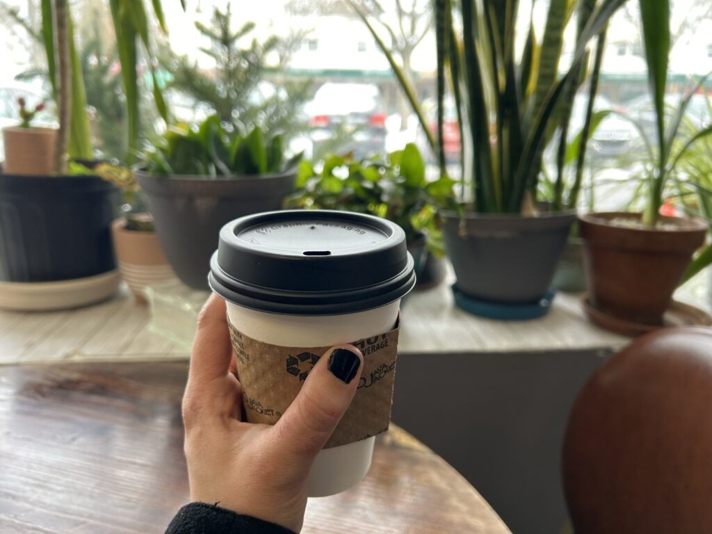 Hand holding up a cup of coffee in front of a table, window, and many potted plants