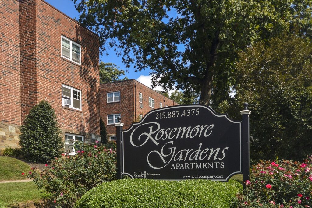A black sign reading "Rosemore Gardens Apartments" in front of several brick buildings and shrubbery