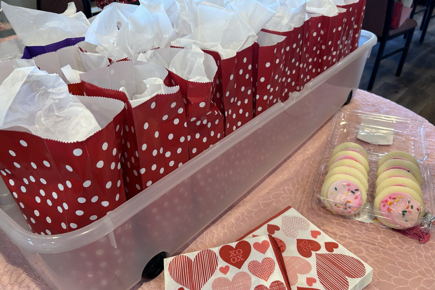 Red goodie bags with white polka dots and white tissue paper sitting on a pink table with cookies and decorative napkins