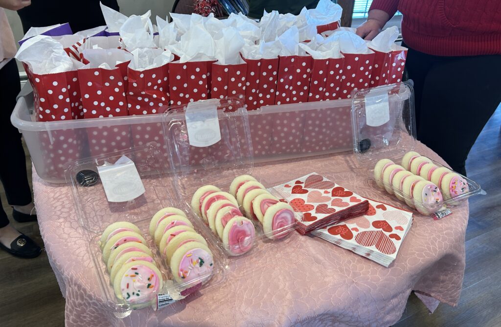 Three boxes of pink frosted sugar cookies on a decorated table in front of a box of red gift bags