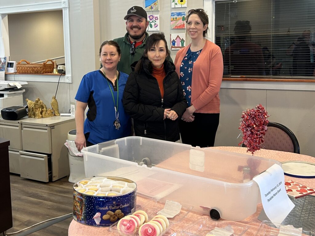 Four people, three women and a man, standing and smiling for a picture inside of an assisted living home. Cookies and decorations are sitting on the table in front of them.