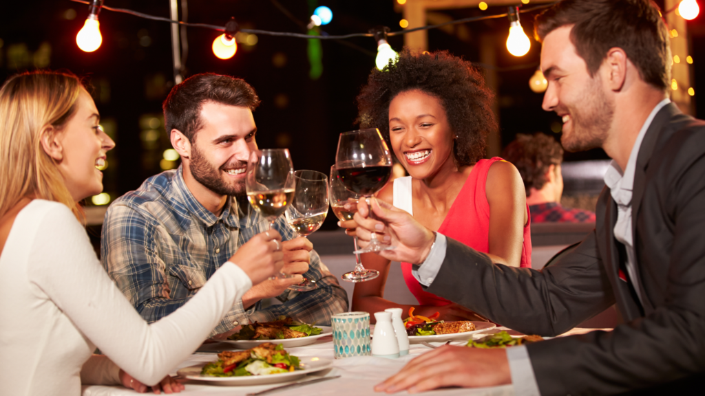 A group of 4 friends dressed nicely and smiling at each other while clinking wine glasses and eating dinner.