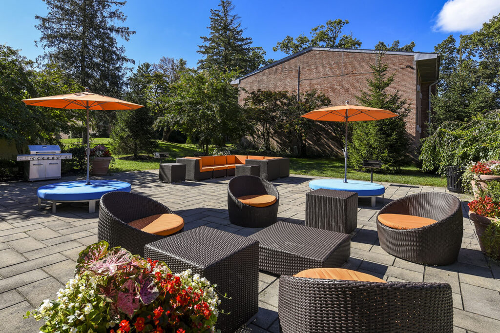 A lounge area with several black wicker tables, chairs, and couches. Orange  cushions on the seats and orange umbrellas connected to the seats. There is a silver gas grill in the shade of a tree.