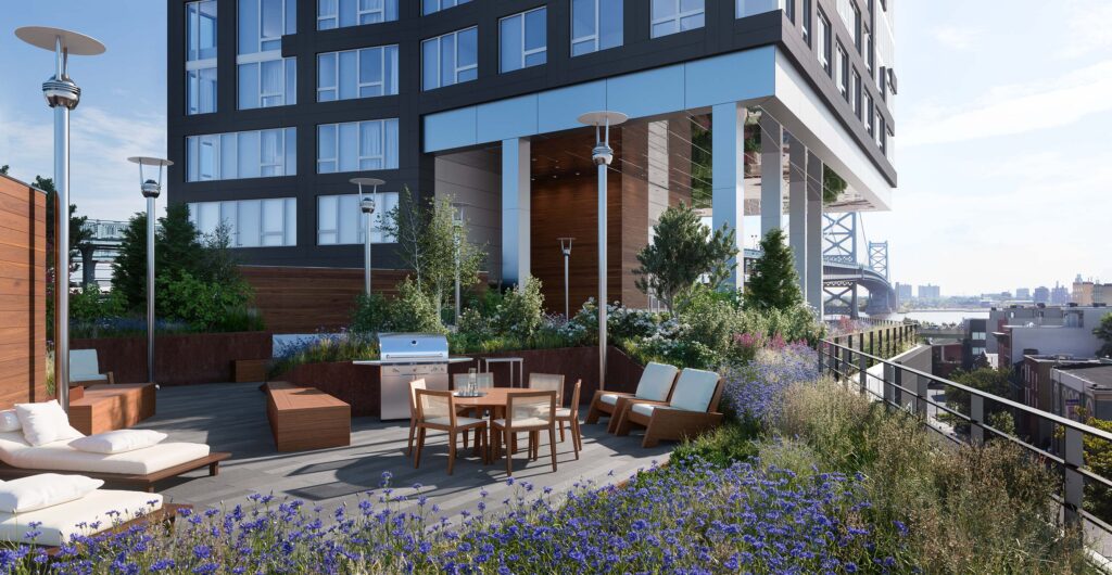 Garden Terrace outside of Bridge. Table, lounge chairs, and a grill with lights and heaters. Surrounded by flowers and greenery. View of the Ben Franklin Bridge.