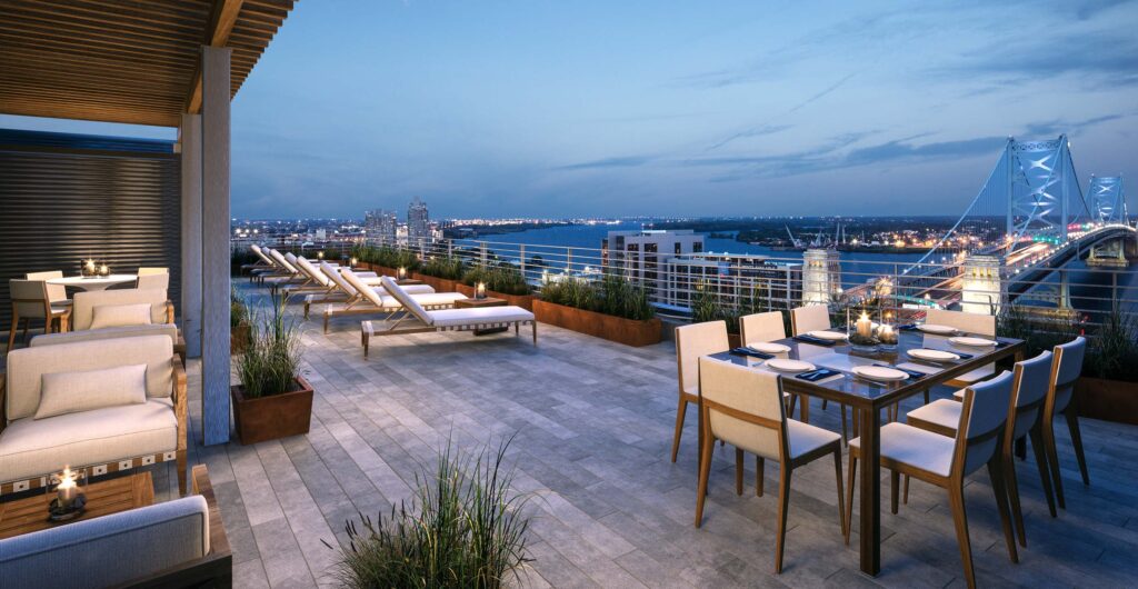 Beautiful rooftop terrace with several tables and chairs, lounge chairs, and greenery. Overlooking the Ben Franklin bridge at dawn, city lights in the distance