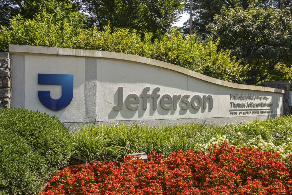 A stone sign with the Thomas Jefferson blue "J" logo in front of green plants and red flowers
