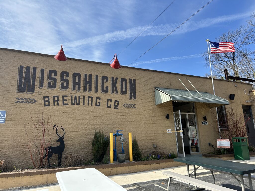 The side of a tan building with large letters that say "Wissahickon Brewing Co" with picnic tables and an American flag flying 