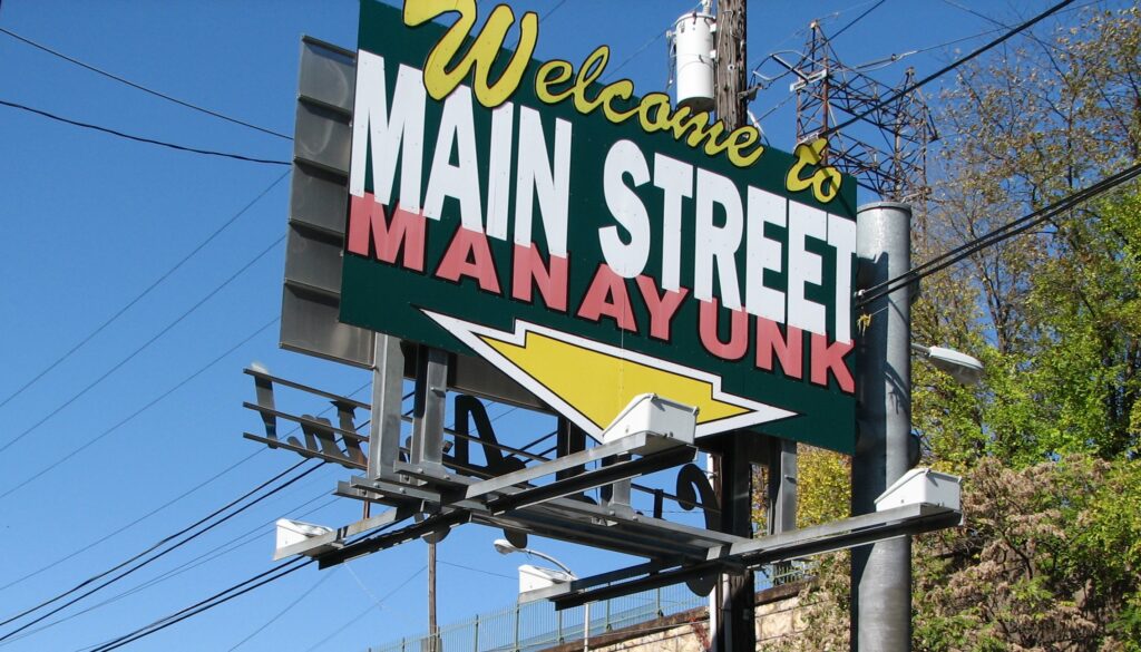 Large street sign with pink, white, and yellow writing that reads, "Welcome to Main Street Manayunk"