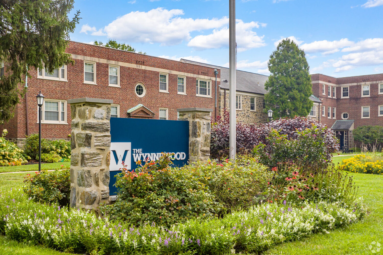 A blue sign with a "W" that reads "The Wynnewood" nestled in some green shrubs and flowers. Tan, brick building in the background