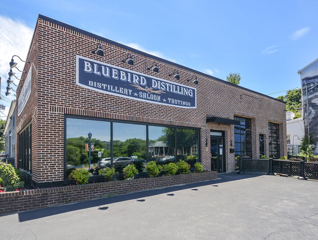 Brick building with greenery in planters. Painted on sign that reads "Bluebird Distilling" with the words "Distillery, Saloon, Tasting" written underneath