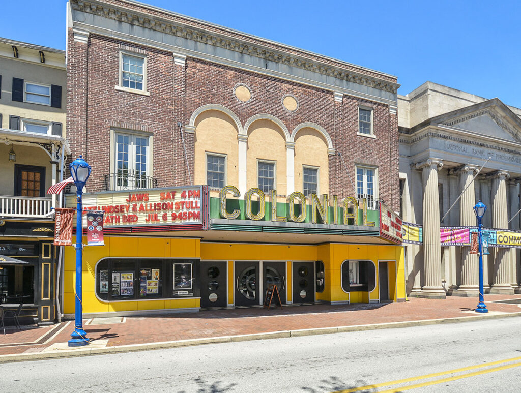 A large, old brick building with a traditional movie theater look to the front. In glowing lights, it says "Colonial" and the shows are listed in red on the side
