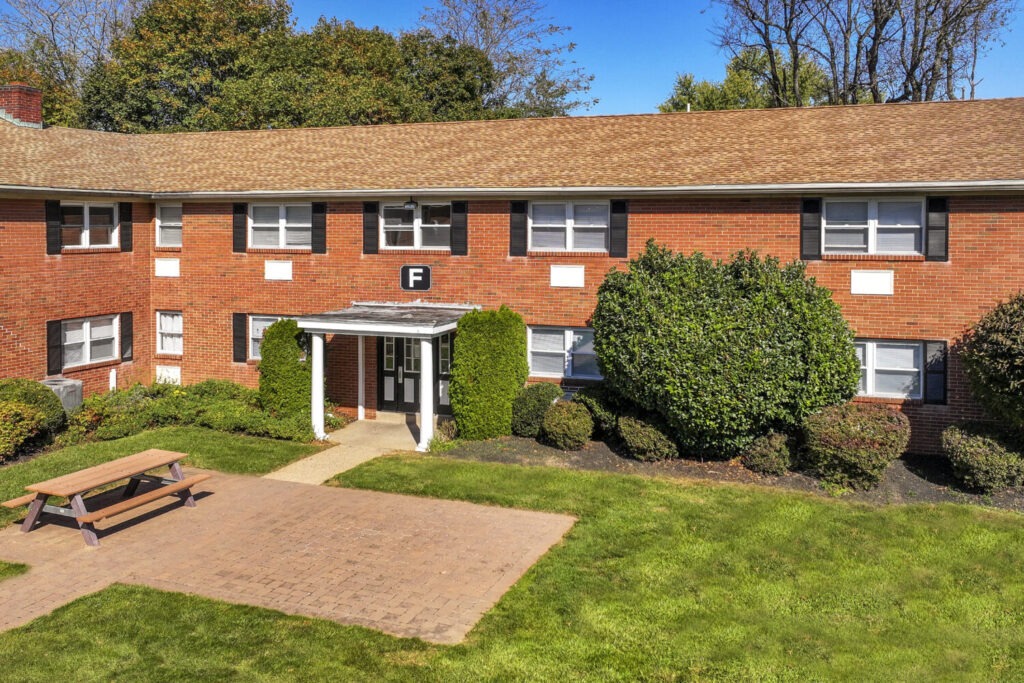 Brick building with a picnic table out front. Black shudders on the windows and large bushes and trees in front.