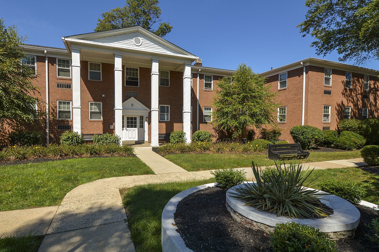 The front of Fair Oaks' Building B, tan brick with white pillars and many green trees and bushes