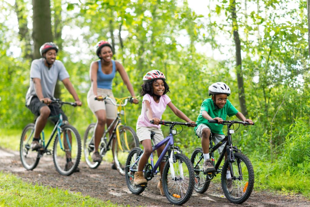 Two parents and two children with helmets on riding bikes on a wooded trail. All smiling and happy.