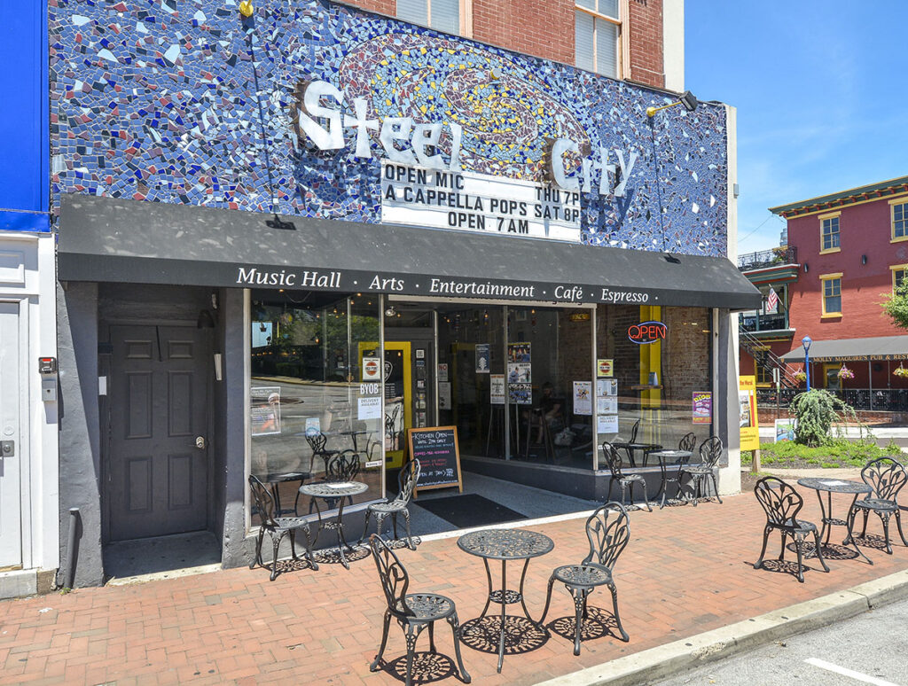 Building with beautiful blue mural reading "Steel City" with a list of upcoming musicians and open mic events. Tables and chairs out front.