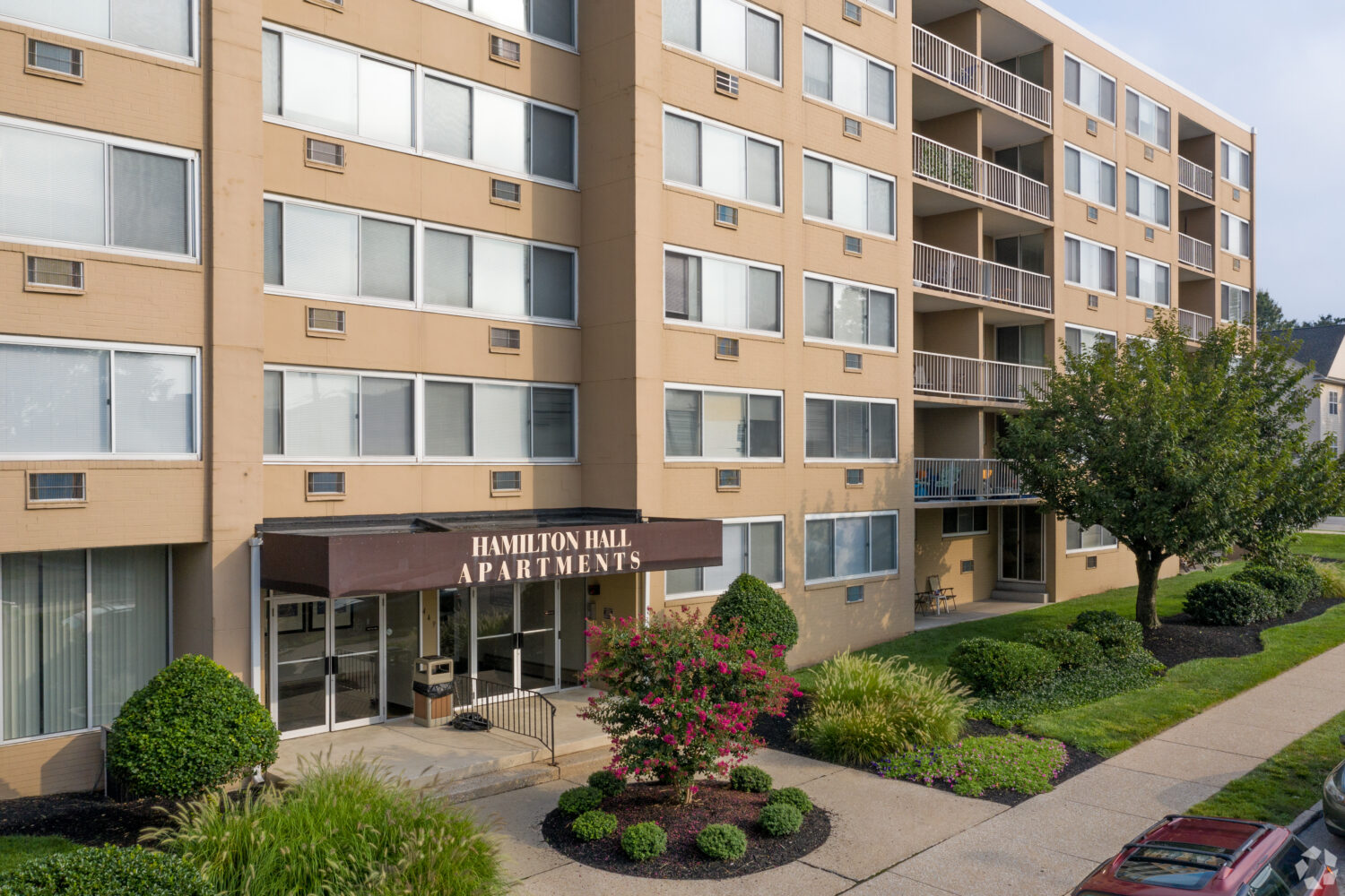 Tan building with a brown awning that reads "Hamilton Hall Apartments" surrounded by trees and bushes