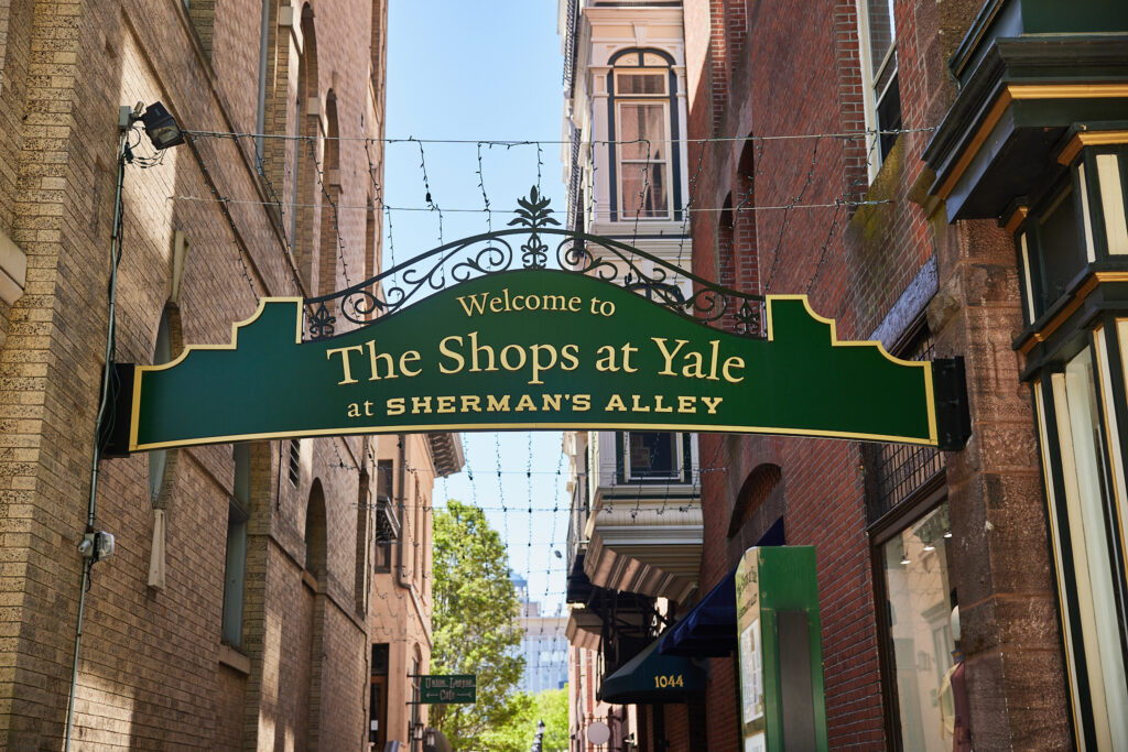 Green ornamental sign that reads "Welcome to The Shops at Yale at Sherman's Alley"