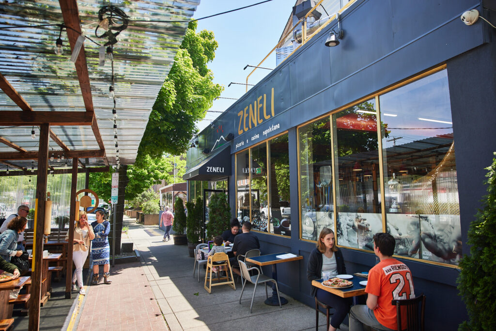 Black painted restaurant with gold paint that reads "Zeneli" with outdoor sidewalk seating with dozens of people sitting in the sun