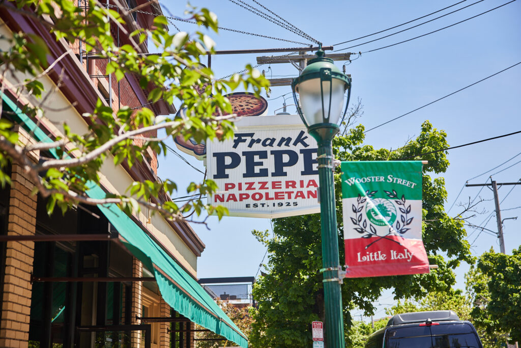 White sign in front of a building reading "Frank Pepe" with green trees in front of it. Next to the sign is a flag with the Italian colors that reads "Wooster Street Little Italy"