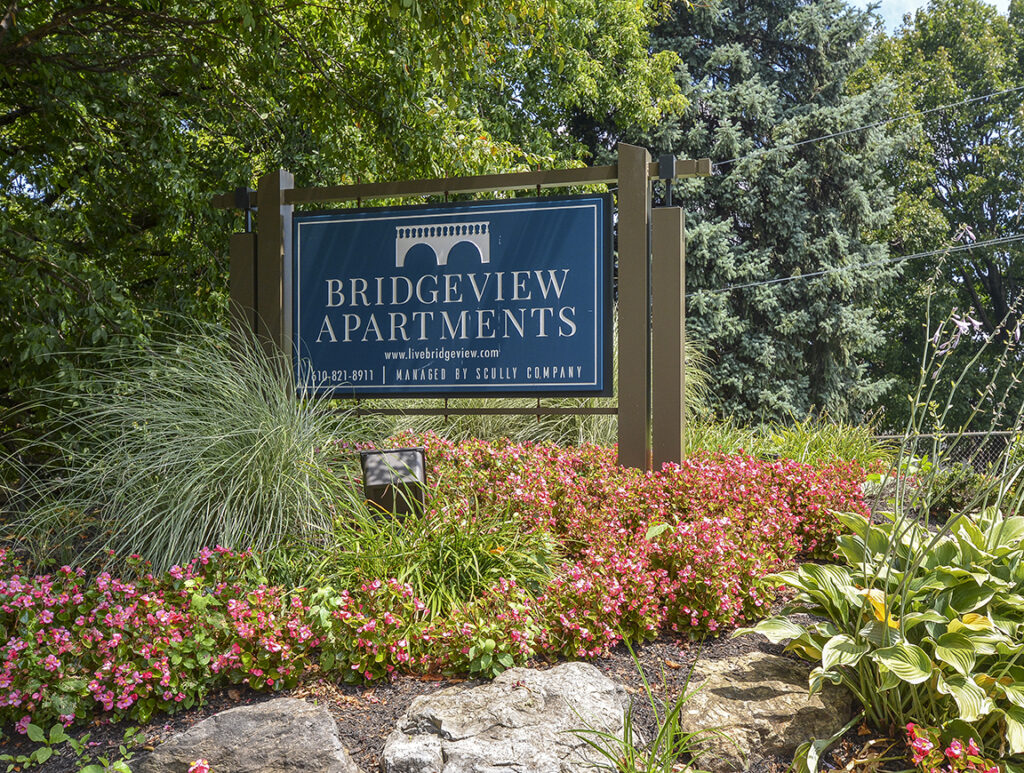 Blue monument sign that reads "Bridgeview Apartments" surrounded by trees and flowers