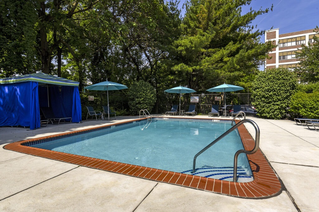 Outdoor pool surrounded by trees with cabanas, tables, chairs, and umbrellas.